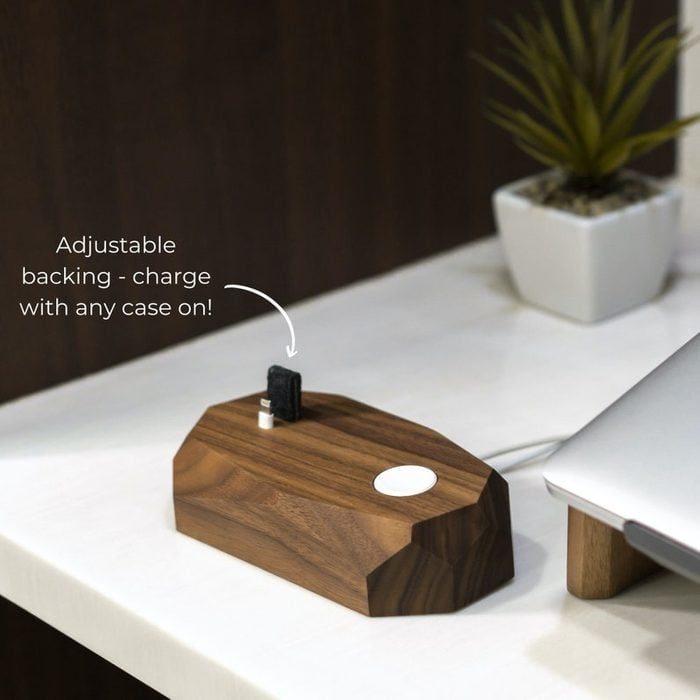Wooden phone dock with adjustable backing sits on the desk, next to a laptop and plant. Text reads, 