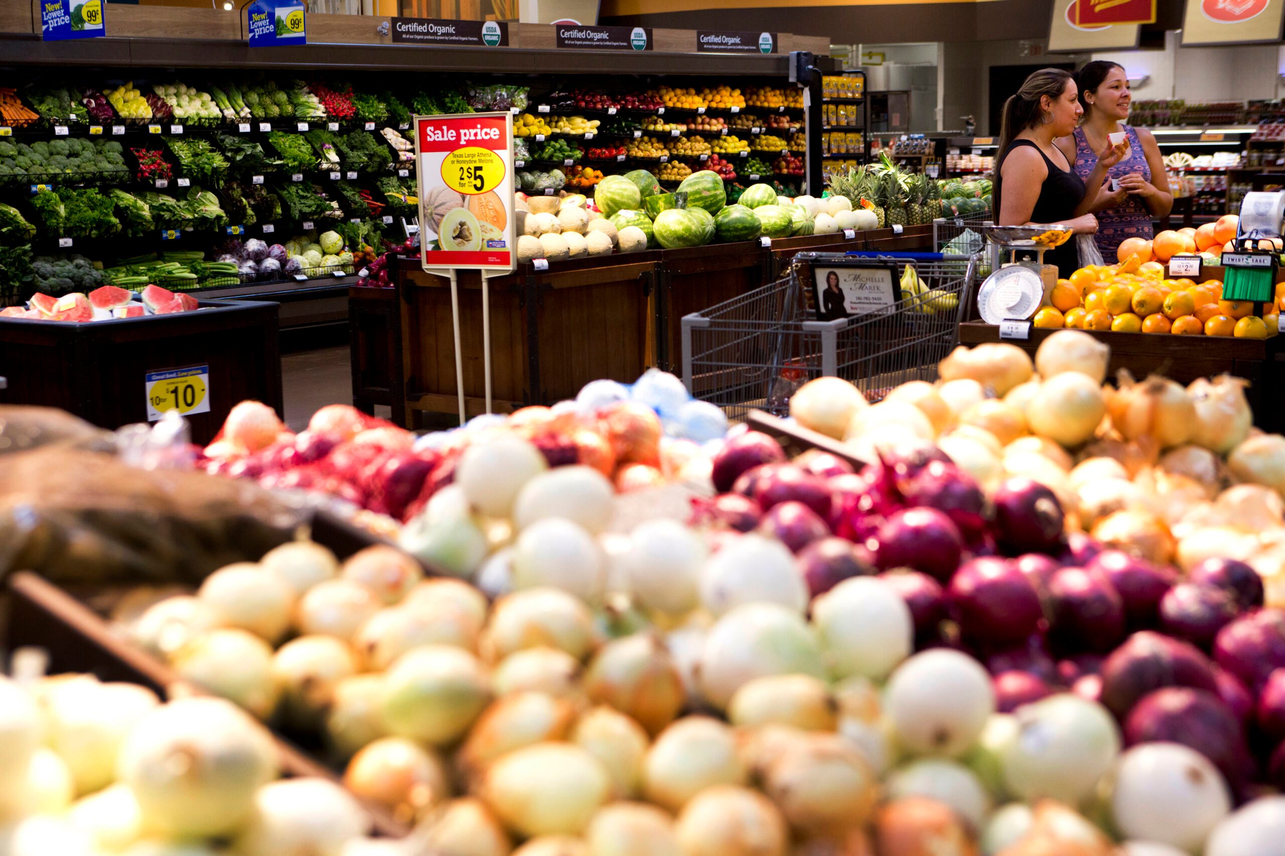 Onions displayed on tables, foreground; two people shopping, background; context is a supermarket produce section. Sign reads: "Sale price, Extra Large Limes, 2 for $5".