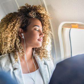 Person wearing earphones relaxes with eyes closed in an airplane seat, sunlight streaming through the window.