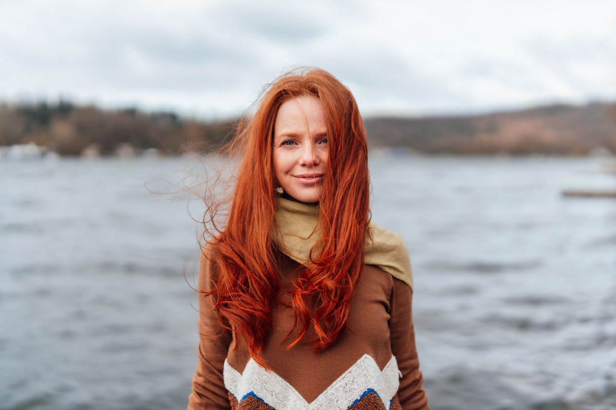 Smiling Redhead Woman With Long Hair In Front Of Lake