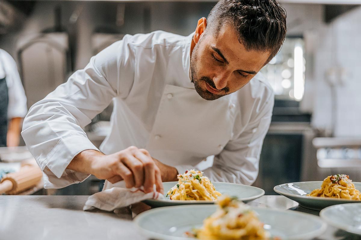 Young male chef cleaning plate of Fettuccine pasta at kitchen counter in restaurant
